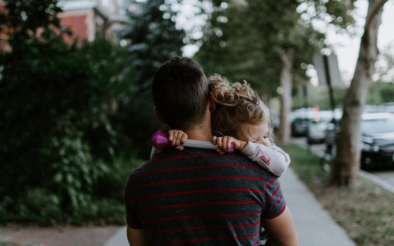 tired little girl rests her head on her father's shoulder as he carries her down the city sidewalk